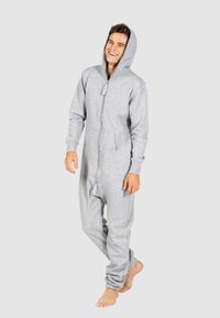 Grijze fleece onesie met een volledige rits, capuchon en voorzakken. Mouwen zijn iets elastisch. Model staat blootvoets tegen een neutrale achtergrond.