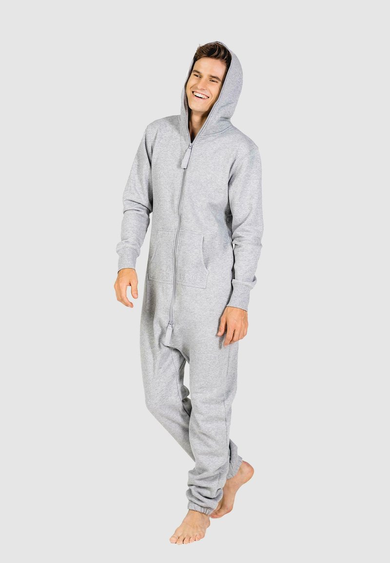 Grijze fleece onesie met een volledige rits, capuchon en voorzakken. Mouwen zijn iets elastisch. Model staat blootvoets tegen een neutrale achtergrond.