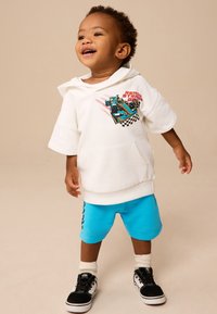 Baltas gobtuvas džemperis su lenktynių grafika, derinamas su mėlynais šortais, kuriuose yra languotos šoninės detalės, ir juodais sportbačiais su baltais pado elementais.