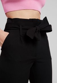 Černé kalhoty na míru mají široký pas a mašličku vpředu, doplněné hlubokými kapsami a hladkou texturou.