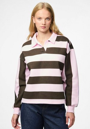 Jonge vrouw in een roze en donkerbruine horizontaal gestreepte longsleeve polo en donkerblauwe jeans, staand voor een witte achtergrond.