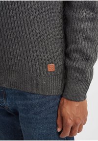 Grå strikket genser med vertikale ribber, utstyrt med en brun lærlapp med "Blend"-merke nær sømmen. Brukt over blå denim.