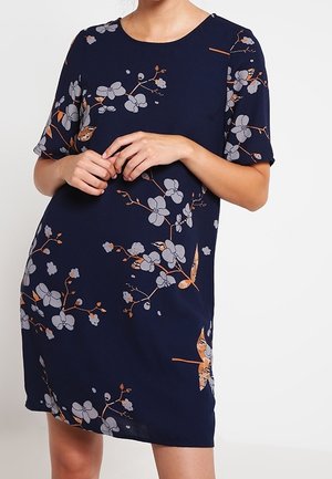 Kvinde iført en mørkeblå kortærmet kjole med lysegrå og orange blomsterprint, stående foran en ensfarvet lys baggrund.