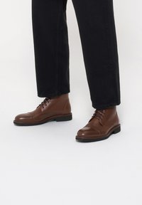 Brązowe skórzane botki za kostkę o gładkim wykończeniu, okrągłym nosku i sznurowanym wzorze. Czarne spodnie zakrywają część butów, podkreślając obuwie.