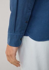 Primo piano del braccio sinistro di una persona che indossa una camicia blu a maniche lunghe con polsini abbottonati e pantaloni bianchi su uno sfondo neutro.