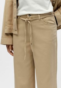Bēša kokvilnas bikses ar auklu pie jostas, vieglu tekstūru, plašām kabatām un daļēji redzamu atbilstošu bēša krāsas žaketi.