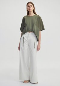 Olivově zelený cropped top s texturovanými rukávy, vybavený stahovací šňůrkou na spodním lemu. V kombinaci s širokými bílými kalhotami a béžovými sandály.