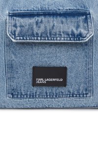 Bolso de ganga azul com aba e etiqueta preta com a inscrição "KARL LAGERFELD JEANS" cosida abaixo da aba no tecido.