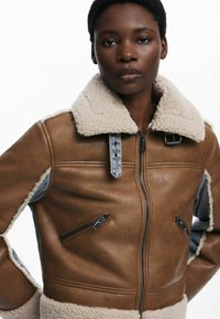 Hnedá kožená bunda so shearling golierom, s šedými denimovými akcentmi a viacerými zipsovými vreckami. Hladká textúra s prispôsobeným strihom.