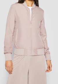 Roze bomberjack met een ritssluiting aan de voorkant, geribde manchetten en zoom, twee zijzakken en een gladde stoftextuur. Gestyled met een wit topje.