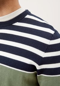Nahaufnahme einer Person, die einen gestreiften Pullover in Marineblau, Weiß und Grün trägt, wobei ein weißes Unterhemd am Halsausschnitt sichtbar ist.
