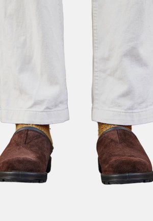 Scarpe slip-on in suede marrone indossate con calzini color senape e pantaloni bianchi visibili dalla parte inferiore dei polpacci.