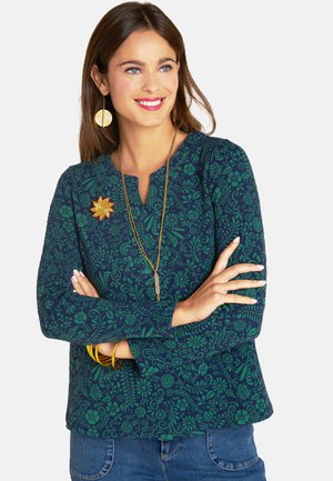 Langärmliges Oberteil in Marineblau mit grünem Blumenmuster, featuring einem Rundhalsausschnitt, einer goldenen Brosche und einer entspannten Passform. Kombiniert mit Jeans.