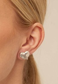 Pendiente de forma de corazón plateado con superficie pulida, ajustado al oído. Diseño simple que destaca la textura suave y la calidad reflectante.