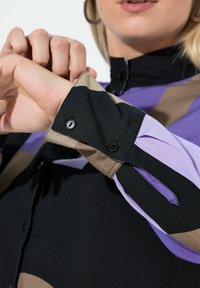 Vrouw die de geruite manchetknoop van een kleurvlakkenoverhemd met zwarte, paarse en beige strepen verstelt.