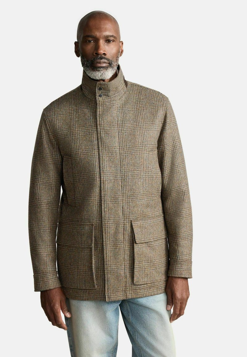 Pruunist ruudulisest jakist kõrge kraega, ees olevate taskutega ja tekstuuriga viimistlusega. Kanda helesiniste sirgete lõikega teksade peal.