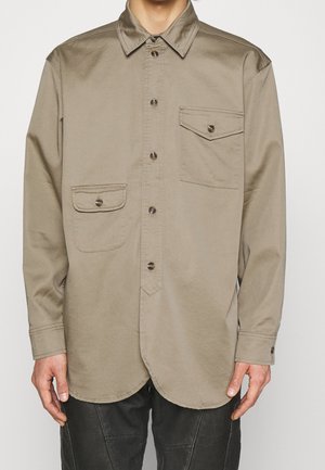 Mand iført en beige skjorte med knapper, to frontlommer med klapper og lange ærmer, kombineret med sorte bukser.