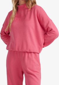 Felpa rosa con alto colletto e scollatura a coste. Realizzata in tessuto morbido, presenta spalle scese e un logo ricamato sul petto.