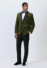 Mees, kes kannab tumerohelist mustrilist sametist smokki, musta liblikat, valget särki, musti pükse ja musti kingi, seisab käed taskutes.