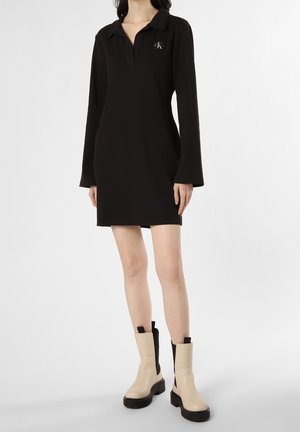 Femme portant une robe mini noire à manches longues avec un col et des bottines beige avec des côtés élastiques noirs.