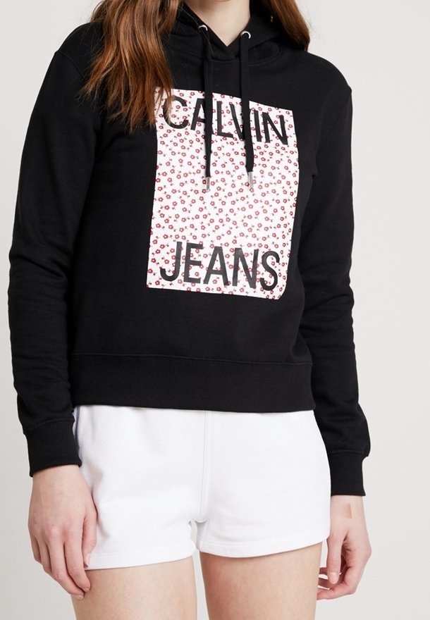 Femme portant un sweat à capuche Calvin Jeans noir avec un patch logo floral et un short blanc, debout devant un fond uni.