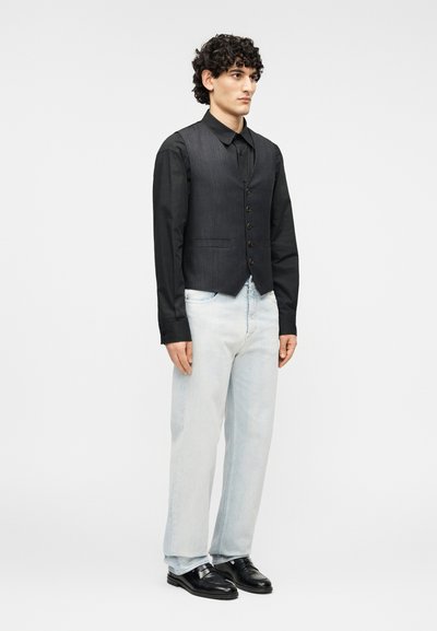 Γκρι γιλεκό με ρίγες πάνω από μαύρο πουκάμισο, σε συνδυασμό με ανοιχτού μπλε τζιν και μαύρα δερμάτινα loafers. Το μοντέλο στέκεται μπροστά σε απλό φόντο.