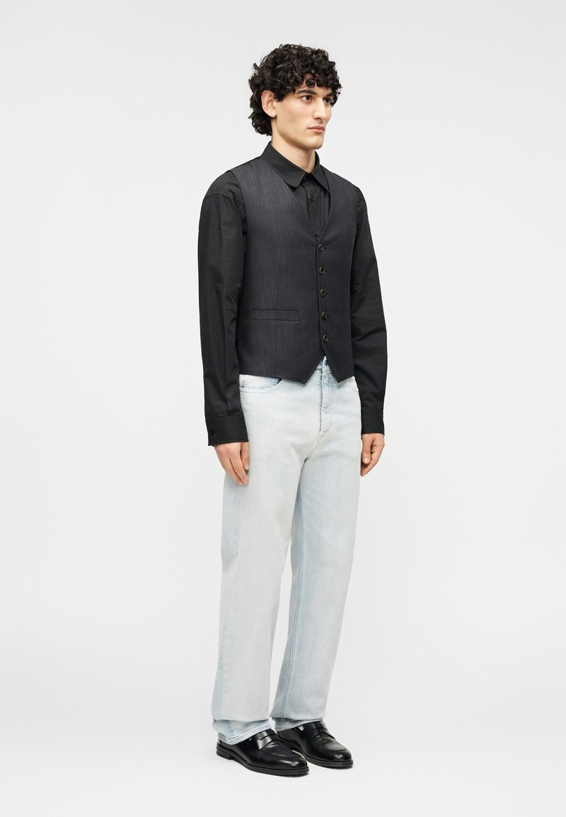 Gilet gris à rayures fines porté sur une chemise noire, associé à un jean bleu clair et des mocassins en cuir noir. Le mannequin est debout devant un fond uni.