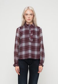 Pikkade blondide juustega naine kannab läbipaistvat burgundi ja halli ruudulist pluusi ripsitud nööbisahtliga ja tumedate teksadega, seistes valge taustaga.