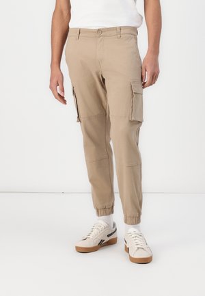 Beige cargo-housut, joissa on joustavat henkäykset, sivutaskut ja nappikiinnitys, valmistettu kestävästä kankaasta. Yhdistetty vaaleanvärisiin lenkkitossuihin.