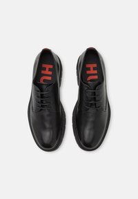 Chaussures en cuir noir à texture lisse, bouts ronds et lacets, avec un logo rouge contrastant sur les semelles intérieures. Semelles plates en caoutchouc.