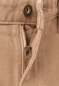 Tissu en denim beige avec une fermeture éclair argentée visible, équipée d'un curseur YKK. Bouton en laiton avec le nom de la marque gravé au-dessus de la fermeture éclair.