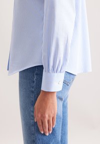 Camicia a maniche lunghe a righe blu e bianche con polsini abbottonati e orlo curvo, abbinata a jeans di denim azzurri, che mostrano una vestibilità rilassata.
