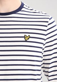 Gestreiftes T-Shirt mit navyblauen und weißen Horizontalstreifen, kurzen Ärmeln und rundem Ausschnitt. Mit gelbem, gesticktem Vogel-Logo.
