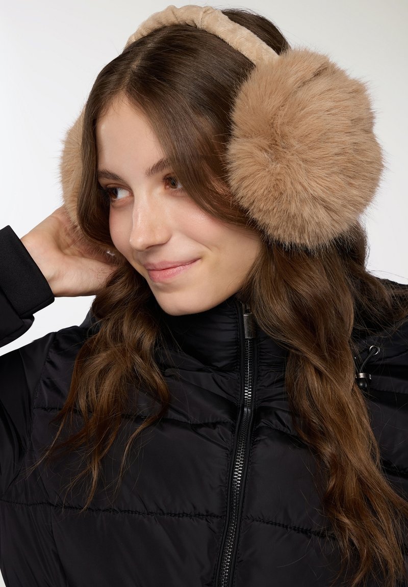 Giovane donna che indossa delle cuffie di pelliccia beige e un giubbotto di piuma nero, con lunghi capelli castani e un leggero sorriso, mentre tocca una cuffia.