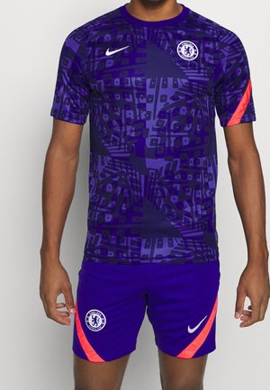 Mężczyzna model ubrany w fioletowy, wzorzysty strój piłkarski Chelsea FC z czerwonymi paskami, herbem klubu oraz białymi logo Nike na koszulce i spodenkach.