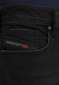 Svarta denimjeans har en bakficka med en röd och svart etikett som har texten "Diesel Industry Denim Division", vilket kompletteras av diskreta sömdetaljer.