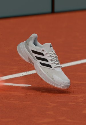 Chaussure de sport blanche et noire soulevant de la terre battue sur un court de tennis en terre battue orange près des marquages de lignes blanches.