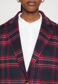 Lähikuva ihmisestä, jolla on päällään tummansininen ja punainen ruudullinen takki valkoisen kauluspuseron päällä, josta näkyy myös helmihelmet.