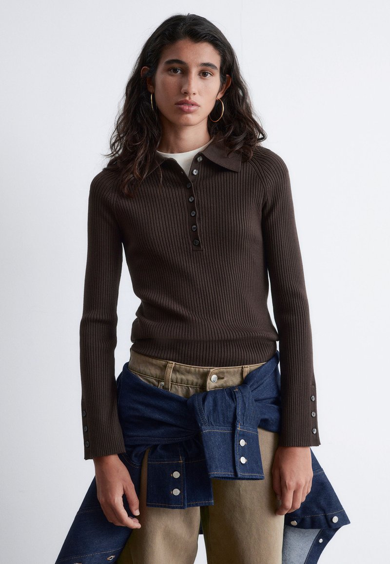 Brown ribbed long-sleeve shirt with a collar, front button closure, and button accents on the sleeves. Worn with denim and khaki pants.