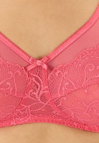 Sostenido de encaje rosa con patrones florales, superposición de material transparente, bordes en forma de ondas y un pequeño lazo en el centro delantero.