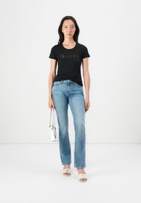 Black fitted t-shirt with embossed "Guess est. 1981" logo, paired with light blue straight-leg jeans, white slides, and a small white handbag.