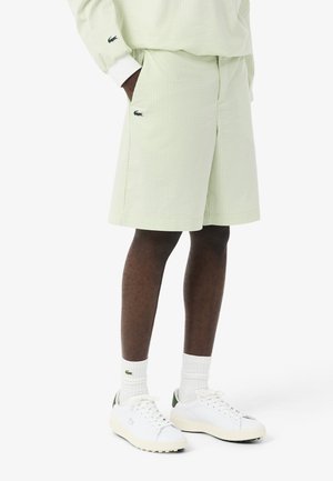 Persona vistiendo pantalones cortos a rayas verde claro y una camiseta de manga larga a juego, con calcetines blancos y zapatillas blancas, todos con el logo de un cocodrilo.