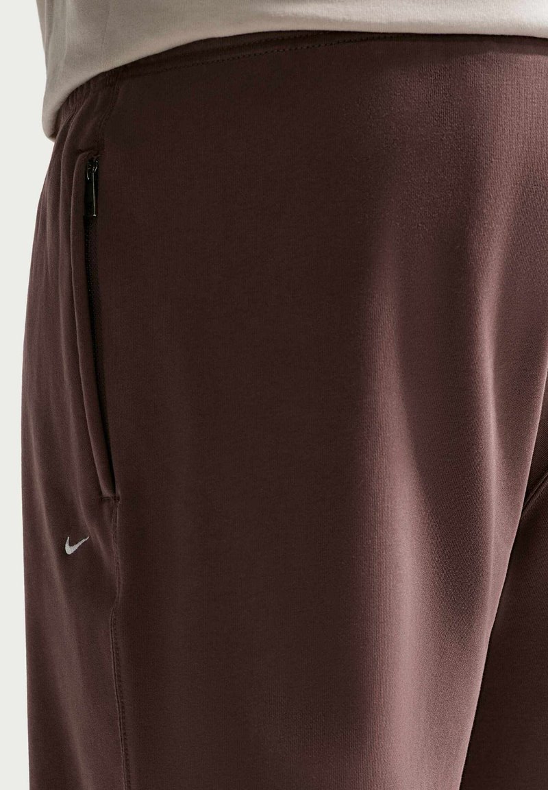 Close-up van een persoon die een bruine Nike-broek draagt met een klein wit Nike-logo en een zijzak met rits, gecombineerd met een licht shirt.