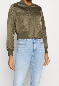 Veste bomber vert olive avec fermeture éclair, deux poches poitrine, poignets et ourlet côtelés. Tissu lisse et brillant avec une coupe structurée.