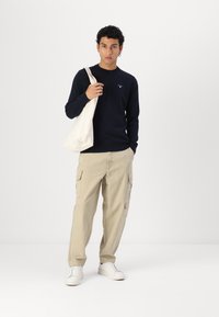 Σκούφος ναυτικού, μπεζ cargo παντελόνια, λευκά sneakers και μια τσάντα tote. Ο σκούφος έχει ένα μικρό λογότυπο, ενώ τα παντελόνια διαθέτουν πολλές πλαϊνές τσέπες.