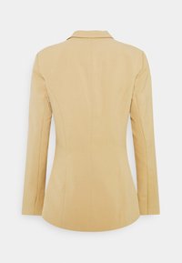 Blazer beige sur mesure avec manches longues, taille cintrée et col plat, vu de dos sur un fond uni.