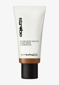 MAC Strobe Dewy Skin Tint dalam tabung putih dengan dasar coklat, dilengkapi teks hitam dan tutup hitam matte. Tekstur yang halus.