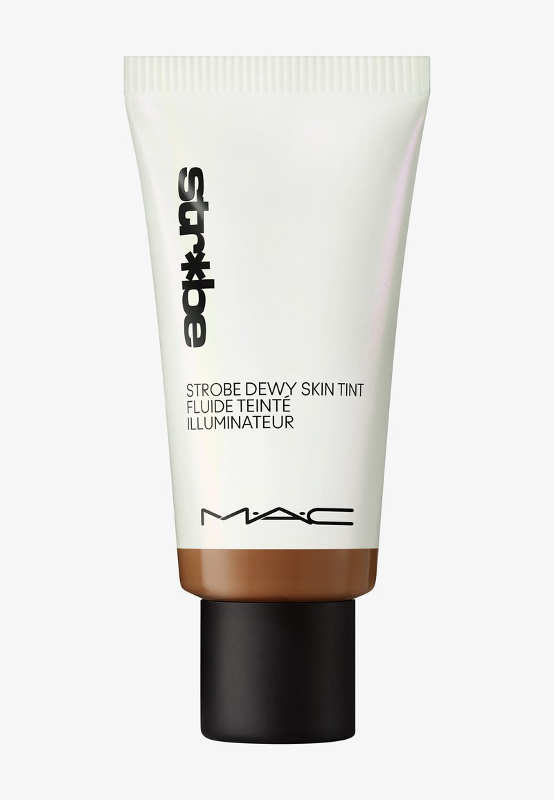 MAC Strobe Dewy Skin Tint dalam tabung putih dengan dasar coklat, dilengkapi teks hitam dan tutup hitam matte. Tekstur yang halus.