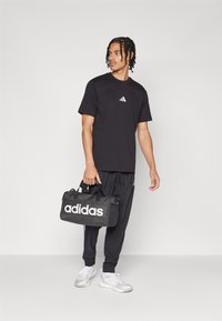 Zwart katoenen T-shirt met wit Adidas-logo, gecombineerd met zwarte broek en een zwarte sporttas gemarkeerd met witte "adidas" tekst. Witte sneakers.