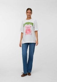 Witte katoenen T-shirt met een roze teddybeer graphic met een strik en "LONDEN" in groene tekst. Gecombineerd met blauwe spijkerbroeken.